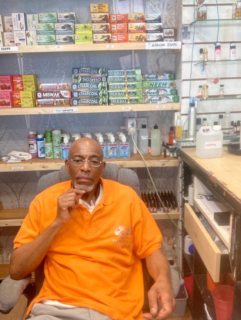Man in an orange shirt sitting in a store with shelves of products.